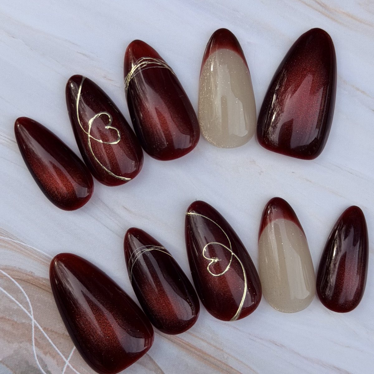 Ten almond press-on nails in burgundy cat-eye, two nude with burgundy tips, with gold heart line art