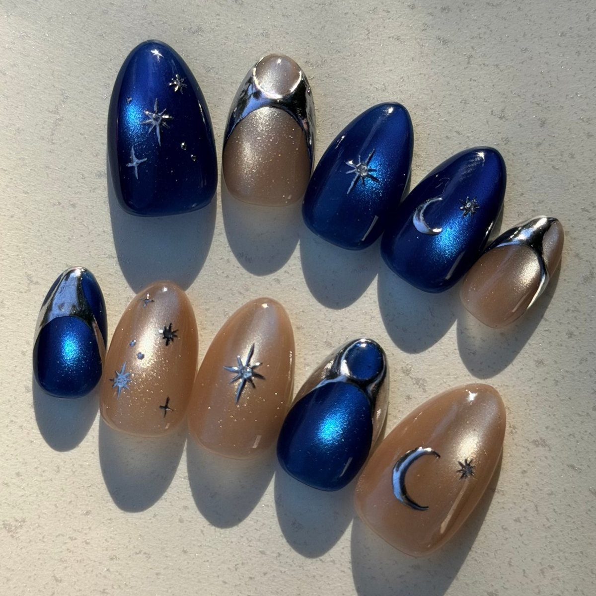 Ten medium almond press-on nails: navy blue cat-eye and nude shimmer with silver chrome French tips, moon and star charms